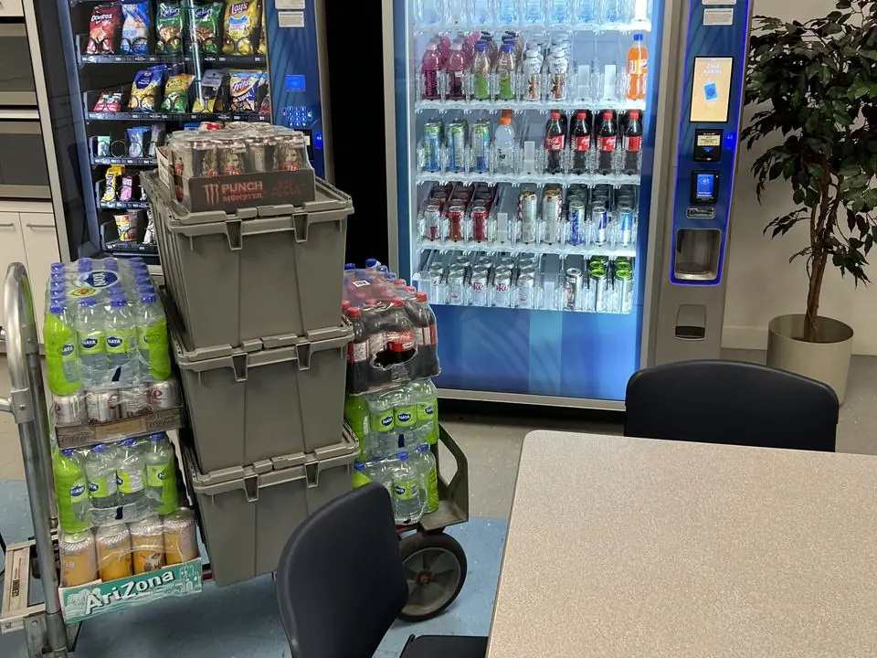 A cart of drink cases and restocking bins next to stocked snack and drink machines in a breakroom