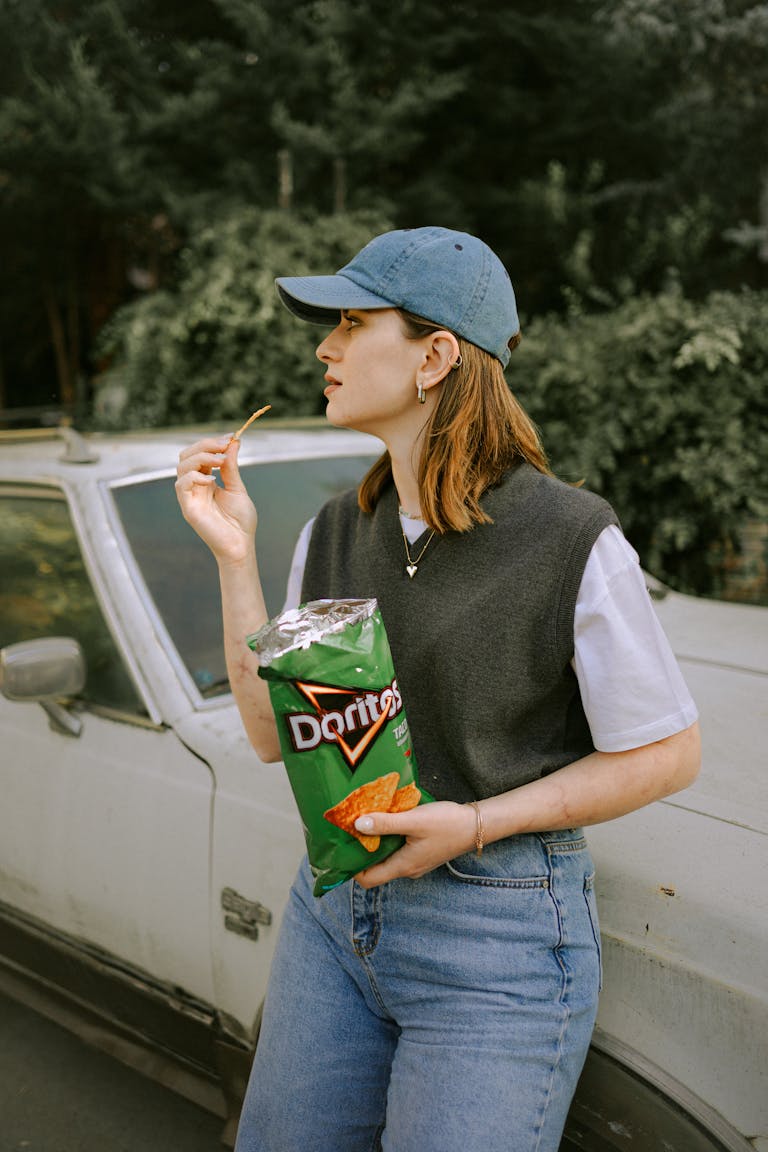 Woman standing by a vintage car, holding and eating Doritos in an outdoor setting.